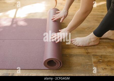 Hohe Winkelansicht der nicht erkennbaren Frau in Sport-Leggings Barfuß auf Holzboden im Wohnzimmer stehen und verdrehen Matte nach Yoga-Übung Stockfoto