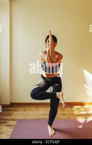 Weibliche Athletin im Sport-Outfit stehen in garudasana Pose während Yoga auf der Matte in der Wohnung am sonnigen Tag üben Stockfoto