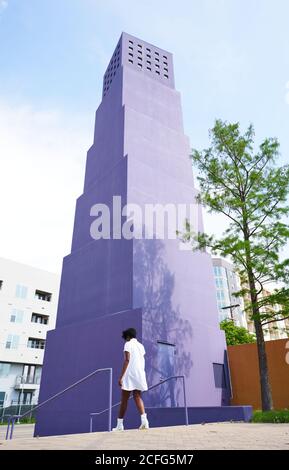 Von hinten betrachtet, wie eine schlanke afroamerikanische Frau in weißem Kleid und Stiefeln allein am sonnigen Sommertag gegen ein mehrstöckiges violettes Gebäude in der Stadt läuft Stockfoto