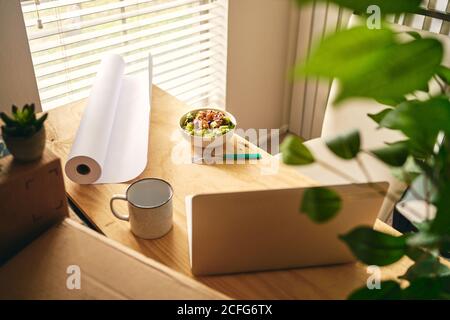 Von oben Holzschreibtisch mit Laptop und Papierrolle Platziert in der Nähe Schüssel mit Salat und Tasse in modernen Wohnung Darstellung der Arbeit von zu Hause Konzept Stockfoto