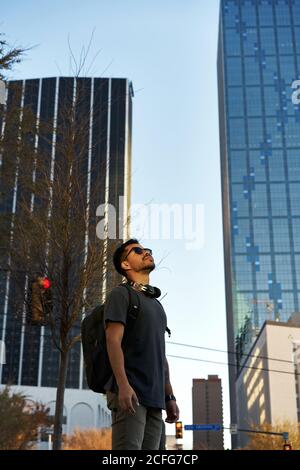 Bärtiger hispanischer Mann im lässigen Outfit lächelnd und aufblickend Mit trendigen Sonnenbrillen und Kopfhörern am Hals in der City Street Stockfoto