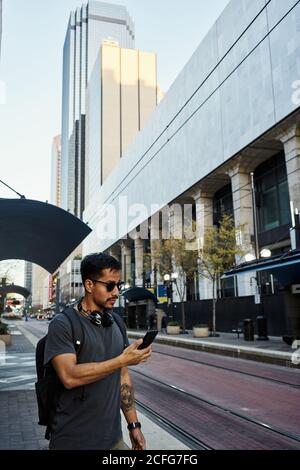 Seitenansicht des zeitgenössischen hispanischen männlichen Touristen in legerer Kleidung Und Sonnenbrillen mit Rucksack und Kopfhörer mit Handy bei Moderne Stadtstraße Stockfoto