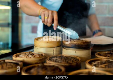 Dim Sum in Bambus Dampfgarer chinesische Küche. Close up Frau weibliche Hände halten Zangen Wahl Mahlzeit köstliche Speisen in Holzkorb. Stockfoto