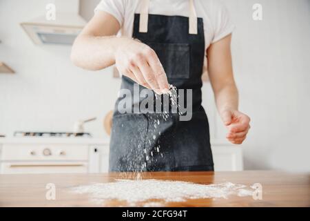 Koch in Schürze streut Mehl mit Hand auf Teig, Holztisch, heller Hintergrund Stockfoto