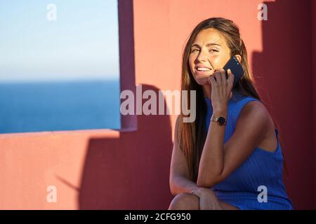 Schöne glückliche Frau, die mit dem Handy auf einer Stufe in einem roten Gebäude sitzt und die Kamera anschaut Stockfoto