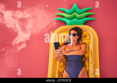 Von oben Frau in Sonnenbrille und Badeanzug auf Ananas Luftmatratze liegend Selfie auf rosa Wasser von Salzsee Stockfoto