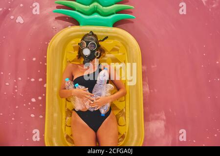 Ansicht von oben auf einer Frau in einem Atemschutzgerät mit Plastikflaschen, die auf einer aufblasbaren Ananas-Matratze im roten Wasser der tropischen Lagune liegen Stockfoto