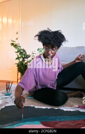 Junge schöne afroamerikanische Frau mit Afro-Haar, Amateur-Künstler im lässigen Outfit Zeichnung buntes Bild mit Pinsel auf Papier, während auf dem Boden in gemütlichen Wohnung sitzen Stockfoto