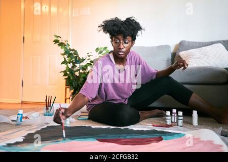 Junge glücklich schöne afroamerikanische Frau, Amateur-Künstler in lässigem Outfit Zeichnung buntes Bild mit Pinsel auf Papier, während auf dem Boden in gemütlichen Wohnung sitzen Stockfoto