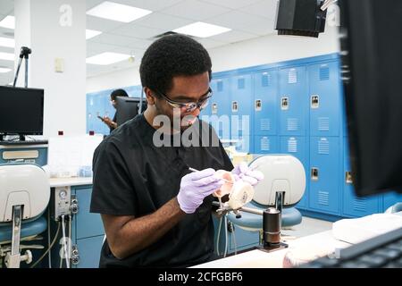 African American Kerl in Latexhandschuhe mit Mund Spiegel und Sonde zur Überprüfung falscher Zähne bei der Arbeit im modernen Labor Stockfoto