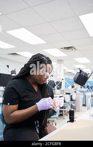 Entzückte afroamerikanische Weibchen mit Zöpfen mit Mundspiegel und Fragen Sie den Mitarbeiter, um falsche Zähne während der Arbeit im Labor zu zeigen Stockfoto