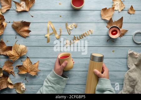 Hände halten isolierte Thermoskanne und Bambusbecher. Null Abfall Tee im Herbst. Flach auf verblassten Holztisch mit vereinzelten Herbstblättern. Text A für Stockfoto