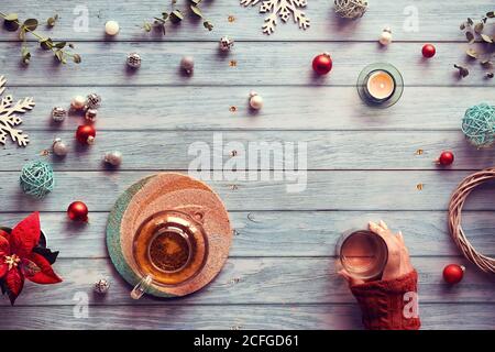 Wintertee, flache Lay-Anordnung mit Glas Teekanne, Glas Tee in der Hand auf verblassten Holzplanken. Weihnachtsdekor - Spiegelkugeln, Kugeln, Spielzeug, ca. Stockfoto