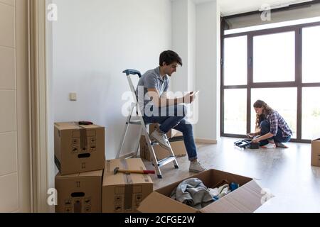 Paar männliche sitzen auf Leiter mit Handy und Frau nimmt Werkzeuge aus der Box in leerer Wohnung mit Karton Boxen Stockfoto