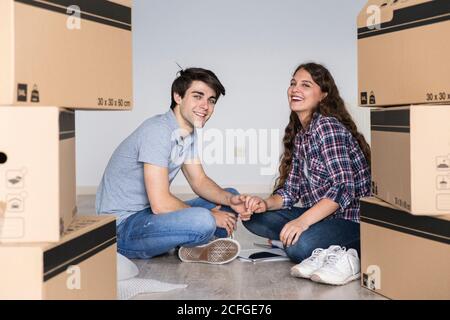 Junges Paar sitzt auf dem Boden mit Kisten halten Hände lächelnd Und Blick auf die Kamera Stockfoto