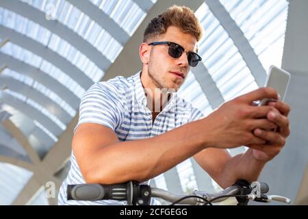 Von unten Schuss von attraktiven jungen Kerl in Sonnenbrille lehnte Am Lenker des Fahrrads und beim Stöbern mit dem Smartphone im überdachten Weg Stockfoto