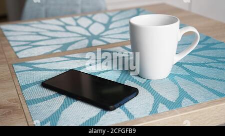 Saubere weiße Tasse mit Griff und Smartphone auf blau Tisch hinter verschwommenem Küchenhintergrund Stockfoto