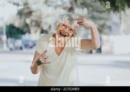 Cool edgy kaukasischen alte Dame Frau mit Schmetterling Tattoo auf der Hand. Schöne funky ältere Frau mit weiß grauen Haaren machen Teufel Rock Zeichen Stockfoto