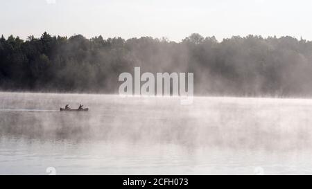 Misty Morning Paddle: Zwei Personen paddeln mit dem Kanu durch den frühen Morgennebel auf einem ruhigen See, während die aufgehende Sonne den aufgehenden Nebel vom warmen See unterstreicht. Stockfoto