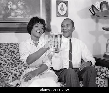 1960 AFROAMERIKANISCHE PAAR MANN FRAU LÄCHELND AUF WOHNZIMMER SITZEN COUCH GENIESSEN TOASTEN MIT PILSNER GLÄSER BIER - N1829 HAR001 HARS 1 COUCH STIL KOMMUNIKATION TEAMARBEIT FREUDE LIFESTYLE ZUFRIEDENHEIT FEIER FRAUEN VERHEIRATET STUDIO SCHUSS EHEGATTEN EHEMÄNNER GESUNDHEIT NACH HAUSE LEBEN KOPIEREN RAUM FREUNDSCHAFT HALBE LÄNGE DAMEN PERSONEN INSPIRATION MÄNNER B&W PARTNER GLÜCK FRÖHLICH GETRÄNK FREIZEIT AFRO-AMERIKANER AFROAMERIKANER AUFREGUNG FLÜSSIGKEIT TOASTEN SCHWARZE ETHNIZITÄT STOLZ GELEGENHEIT LÄCHELT VERBINDUNG PILSNER VERBRAUCHEN HYDRATATION FRÖHLICHE STILVOLLE BREWSKI-ZUSAMMENARBEIT Stockfoto