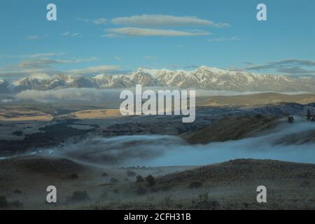 Morgenlandschaft mit einem Flusstal, niedrigen Wolken schleichend auf dem Boden und weißen schneebedeckten Bergen im Hintergrund. Chuya Kamm, Altai Berge, Sibirien, Russland .. Stockfoto