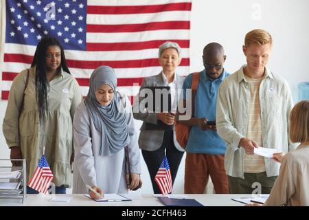 Multiethnische Gruppe von Menschen, die sich in Wahllokalen registrieren, die am Wahltag mit amerikanischen Flaggen geschmückt sind, konzentrieren sich auf die arabische Frau, die im Vordergrund Stimmzettel unterschreibt, Kopierraum Stockfoto