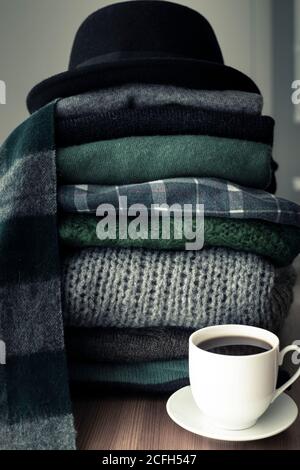 Haufen von grünen und grauen Pullover mit einem schwarzen Hut auf der Oberseite und einem heißen Getränk weißen Tasse auf der Seite, Hintergrund mit Kopierraum, gemütlich, Hygge oder saisonalen Krieg Stockfoto