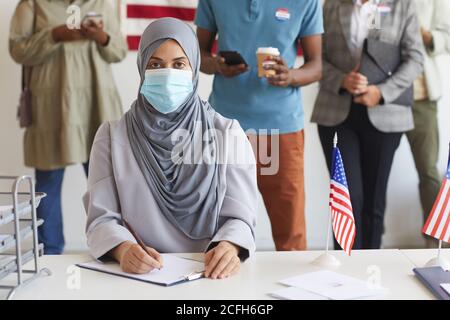 Multiethnische Gruppe von Menschen, die am Wahltag in einer Reihe stehen und Masken in der Wahlstation tragen, konzentrieren sich auf junge arabische Frau, die bei der Registrierung für die Wahl auf die Kamera schaut, Kopierraum Stockfoto
