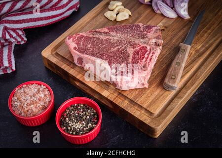 Ein Foto von einem rohen Porterhouse Steak, das auf einem Schneidebrett ruht, überzogen mit Himalaya-Meersalz und frischem gemahlenem Pfeffer, mit Schalotten und Knoblauch i Stockfoto