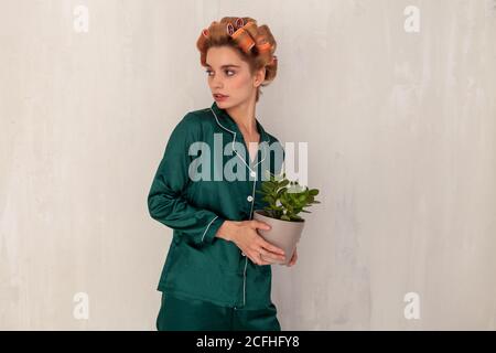 Junge Frau in grüner Seide Pyjamas und Haar Lockenwickler mit Blumentopf in den Händen in der Nähe der strukturierten Wand stehen Stockfoto