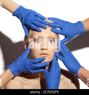 Viele Hände tragen blaue medizinische Handschuhe halten Schönheit weiblich mit Perforationslinien auf Gesicht isoliert auf weißem Hintergrund, plastische Chirurgie Konzept Stockfoto