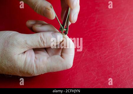 Der Kaukasusmann mittleren Alters schneidet sich die Nägel. Nagelknipser in männlichen Händen auf einem roten Tisch. Gesundheit und Hygiene. Nahaufnahme. Stockfoto