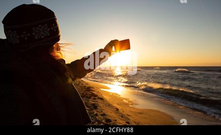 Frau Reisende fotografiert das Meer bei Sonnenaufgang auf einer Smartphone-Kamera. Stockfoto