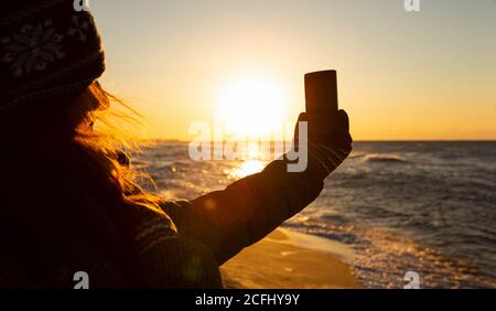 Frau Reisende fotografiert das Meer bei Sonnenaufgang auf einer Smartphone-Kamera. Stockfoto