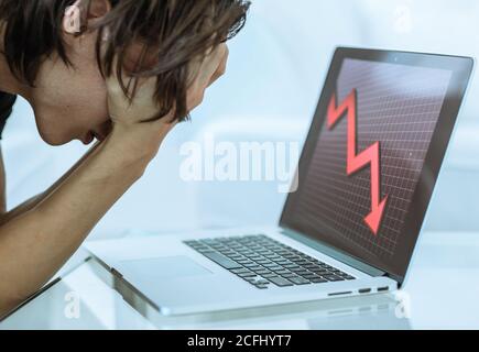 Traurige männliche Aktieninvestor Geld mit Händen auf dem Gesicht zu verlieren, wie der Laptop-Computer zeigt einen Finanzmarktcrash. Wirtschaftlicher roter Pfeil geht nach unten. Stockfoto