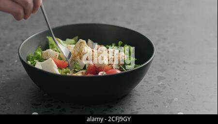 Man handpflücken Salat mit Mozzarella, Kirschtomaten und Friseeblättern in schwarzer Schüssel auf Terrazzooberfläche, breites Foto Stockfoto