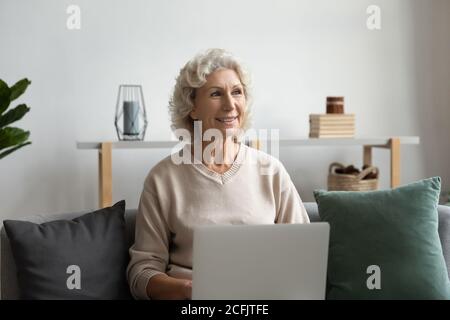 Lächelnd verträumt reife Frau sitzt mit Laptop auf gemütliche Couch Stockfoto