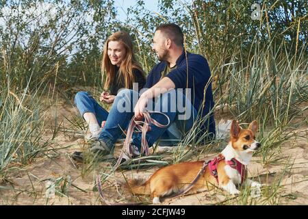 Junge glückliche Paar von Mann und Frau mit Corgi Hund sitzt am Sand. Zwei Personen mit Haustier genießen schönen sonnigen Nachmittag am Strand Stockfoto