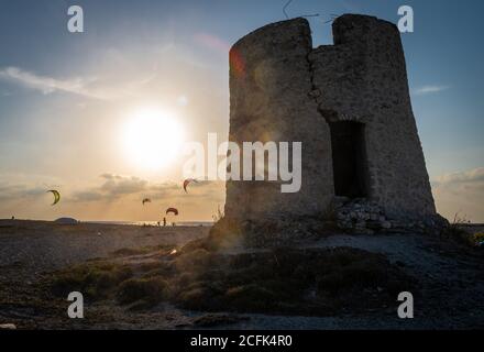 Blick auf den Sonnenuntergang über einer verwesenden Steinwindmühle am Strand von Agios Ioannis mit Kitesurfen im Hintergrund. Stockfoto