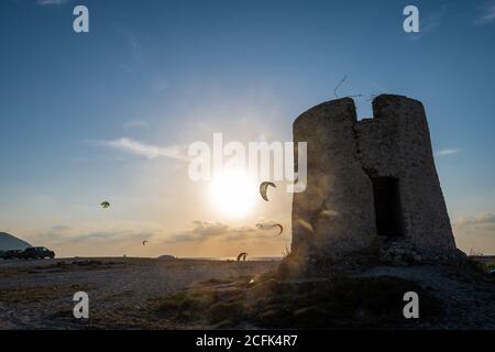 Blick auf den Sonnenuntergang über einer verwesenden Steinwindmühle am Strand von Agios Ioannis mit Kitesurfen im Hintergrund. Stockfoto