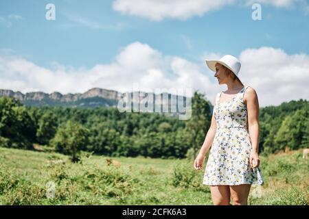 Seitenansicht der ruhigen Frau in Kleid und Hut aufwachen Entlang der Wiese auf dem Hintergrund der Kühe grasen in Bergtal Stockfoto