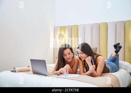 Junge Freundinnen liegen auf dem Bett mit Laptop und lesen Dokumente während der gemeinsamen Vorbereitung auf die Untersuchung Stockfoto