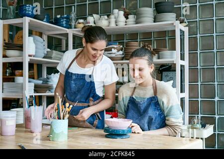 Frau, die Keramik macht. Attraktive qualifizierte junge Dame in Schürze, die am Tisch steht und Schüler unterrichtet. Konzept für Werkstatt und Masterclass Stockfoto