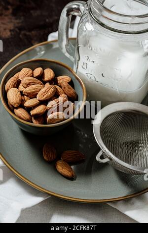 Von oben Keramikschale mit frischen Mandeln auf den Teller gelegt In der Nähe Flasche vegane Milch und Sieb Stockfoto