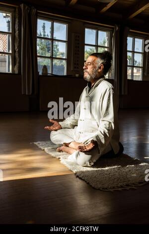 Ganzkörper-Seitenansicht des fokussierten bärtigen ethnischen älteren Mannes Chi-Kung-Meditation in Lotusposition im dunklen Studio üben Stockfoto