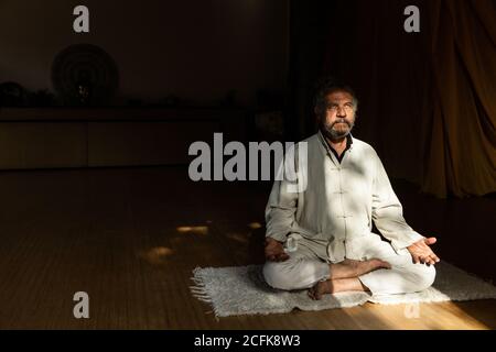 Ganzkörper-Seitenansicht des fokussierten bärtigen ethnischen älteren Mannes Chi-Kung-Meditation in Lotusposition im dunklen Studio üben Stockfoto