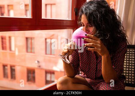 Ethnische nachdenkliche lateinerin genießt einen gesunden Kefir und Pitaya Smoothie, der zu Hause an den Fenstern sitzt Stockfoto