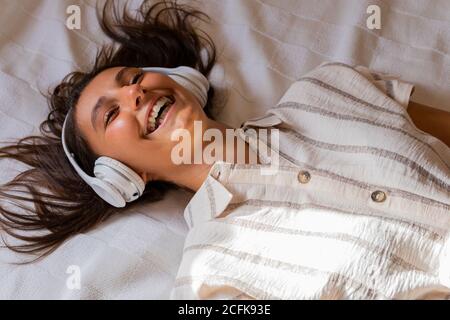 Crop Kind mit Kopfhörern einer Frau, die im Schlafzimmer auf dem Bett liegt und Musik hört, während Sie die Augen schließen Stockfoto