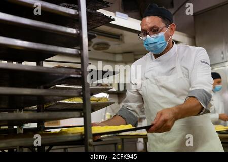 Männlicher Koch trägt Uniform und medizinische Maske Vorbereitung leckere Gebäck In der Küche der Bäckerei während der Coronavirus-Epidemie Stockfoto