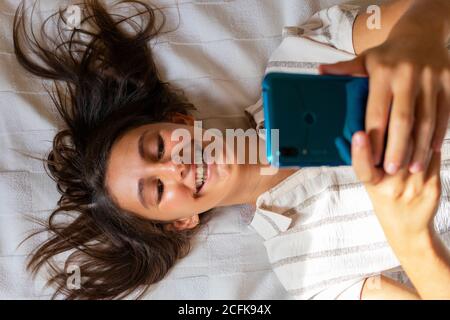 Von oben erfreut Weibchen liegend auf dem weichen Bett bei Home und Lesen von Nachrichten in sozialen Medien über das Mobiltelefon Stockfoto